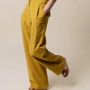 May include: Mustard yellow wide-leg trousers with a high waist and pleats. The trousers have a button closure and side pockets. The model is wearing brown sandals. The trousers are made of a lightweight fabric.