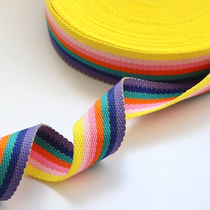 May include: A yellow, pink, orange, green, and purple striped webbing ribbon. The ribbon is rolled up and a section is unrolled, showing the stripes.