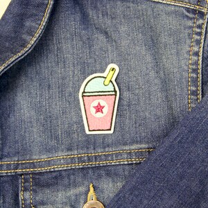 May include: A pink and blue embroidered patch of a slushie with a yellow straw and a pink star on a denim jacket.