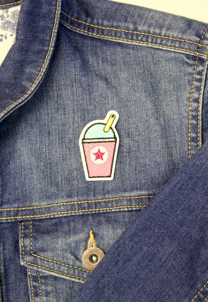 May include: A pink and blue embroidered patch of a slushie with a yellow straw and a pink star on a denim jacket.