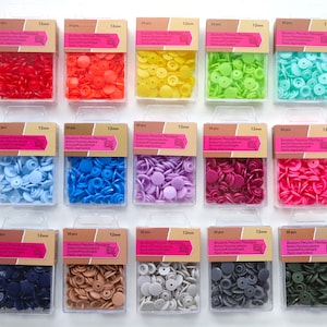 K&ouml;nnte beinhalten: Sortiment von 12 mm gro&szlig;en Druckkn&ouml;pfen aus Kunststoff in verschiedenen Farben, darunter Rot, Orange, Gelb, Gr&uuml;n, Blau, Lila, Rosa, Braun, Grau und Gr&uuml;n. Jede Farbe ist in einem durchsichtigen Plastikbeh&auml;lter mit dem Etikett "Boutons Pression Plastique 12mm" verpackt.