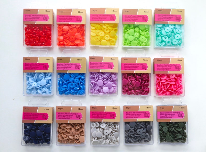 K&ouml;nnte beinhalten: Sortiment von 12 mm gro&szlig;en Druckkn&ouml;pfen aus Kunststoff in verschiedenen Farben, darunter Rot, Orange, Gelb, Gr&uuml;n, Blau, Lila, Rosa, Braun, Grau und Gr&uuml;n. Jede Farbe ist in einem durchsichtigen Plastikbeh&auml;lter mit dem Etikett "Boutons Pression Plastique 12mm" verpackt.
