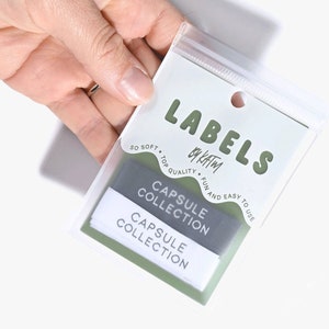 Peut inclure: Un sac en plastique transparent contenant deux étiquettes blanches avec le texte "CAPSULE COLLECTION" en gris et noir. Le sac est muni d'une étiquette blanche sur laquelle est inscrit "LABELS by Katim" en vert. L'étiquette indique également "SO SOFT. TOP QUALITY. FUN AND EASY TO USE".