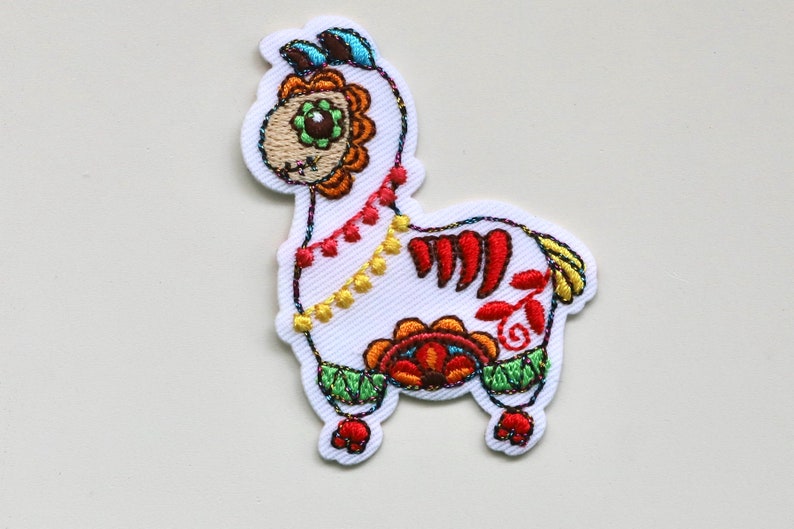 May include: Embroidered white alpaca patch with colourful Day of the Dead inspired details. The alpaca has a floral design on its head and body, with red, orange, yellow, green, and blue accents.