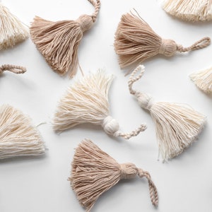 May include: A collection of ten handmade tassels in two colours: beige and white. The tassels are made of soft, fluffy yarn and have a simple, elegant design.