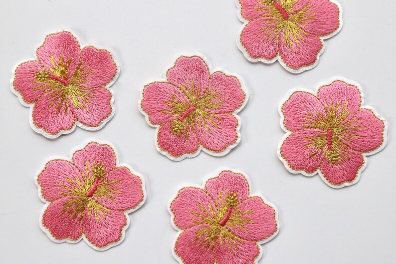 Puede incluir: Seis parches bordados de flores de hibisco rosas y doradas. Las flores tienen un contorno blanco y est&aacute;n sobre un fondo blanco.