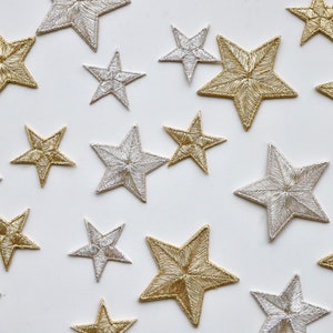 May include: A collection of embroidered star patches in gold and silver, scattered on a white background. The stars are of varying sizes and appear to be made for decorative use, possibly for embellishing garments or accessories.