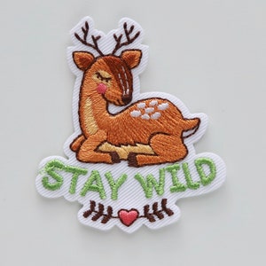 May include: Embroidered iron-on patch featuring a brown deer with white spots, lying down with its eyes closed. The patch has green text that says "STAY WILD" and a small red heart with brown stems.