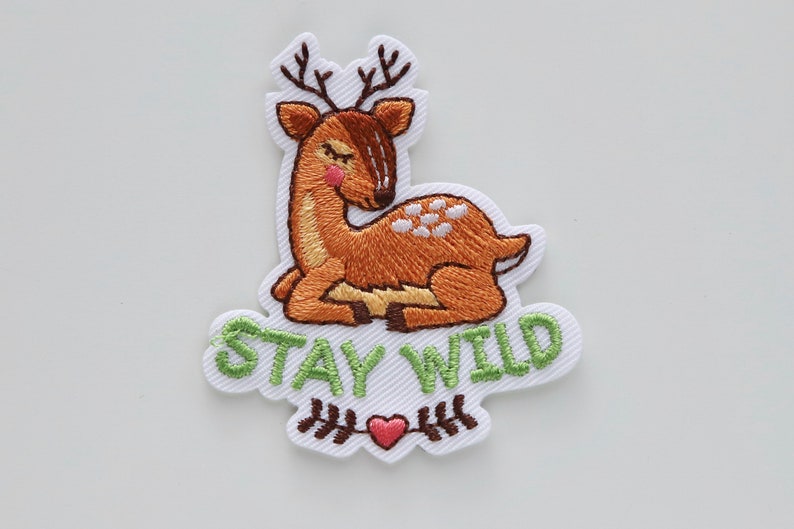 May include: Embroidered iron-on patch featuring a brown deer with white spots, lying down with its eyes closed. The patch has green text that says "STAY WILD" and a small red heart with brown stems.