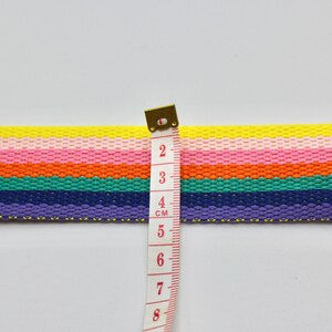 May include: A rainbow striped fabric ribbon with a measuring tape showing 4 cm. The ribbon is made of woven cotton and has a yellow, pink, orange, green, blue, and purple color scheme.