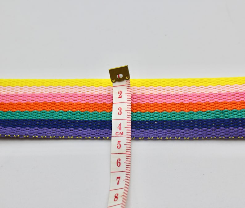 May include: A rainbow striped fabric ribbon with a measuring tape showing 4 cm. The ribbon is made of woven cotton and has a yellow, pink, orange, green, blue, and purple color scheme.
