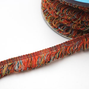 May include: A spool of colorful fringe trim with a mix of orange, yellow, blue, and black threads. The fringe is about 1 inch wide and has a woven base.