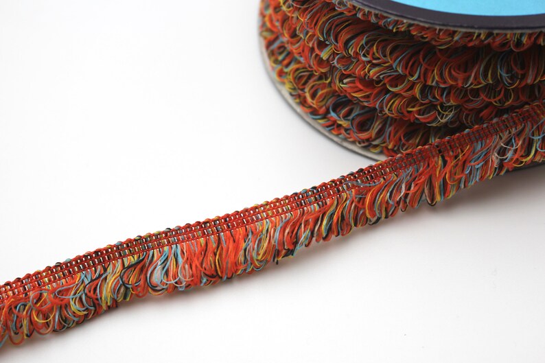 May include: A spool of colorful fringe trim with a mix of orange, yellow, blue, and black threads. The fringe is about 1 inch wide and has a woven base.