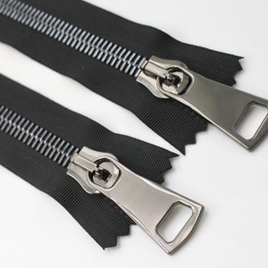 May include: Two black zipper pulls with silver metal sliders and teeth. The zippers are laid out flat on a white surface.