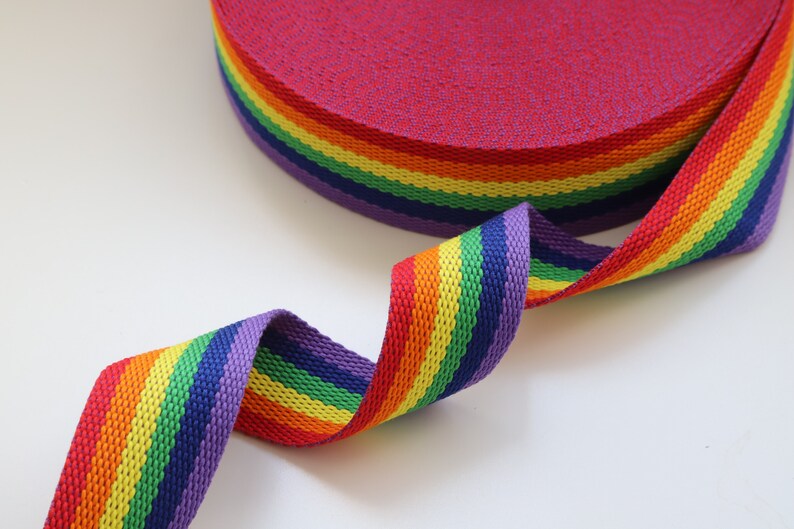 May include: Rainbow striped webbing, a type of flat woven strap, in red, orange, yellow, green, blue, indigo, and violet. The webbing is rolled up and a section is unfurled, showing the colors.