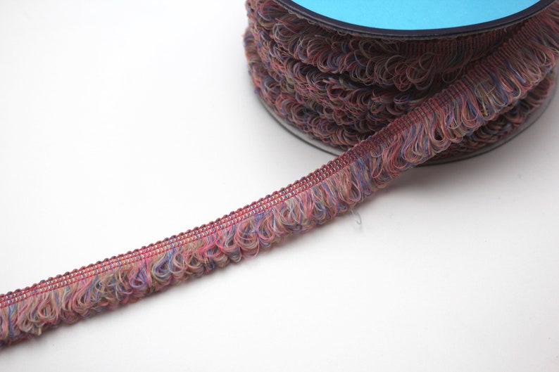 May include: A spool of multi-colored fringe trim with a blue plastic spool. The fringe is a mix of pink, blue, and beige.