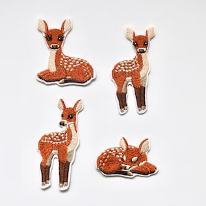May include: Four embroidered iron-on patches of brown and white spotted fawns in different poses. The fawns are all facing forward, with one lying down, one standing, and two sitting.