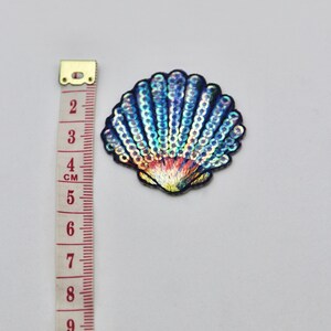 May include: A blue and iridescent seashell-shaped patch with sequins. The patch is about 4 cm wide.