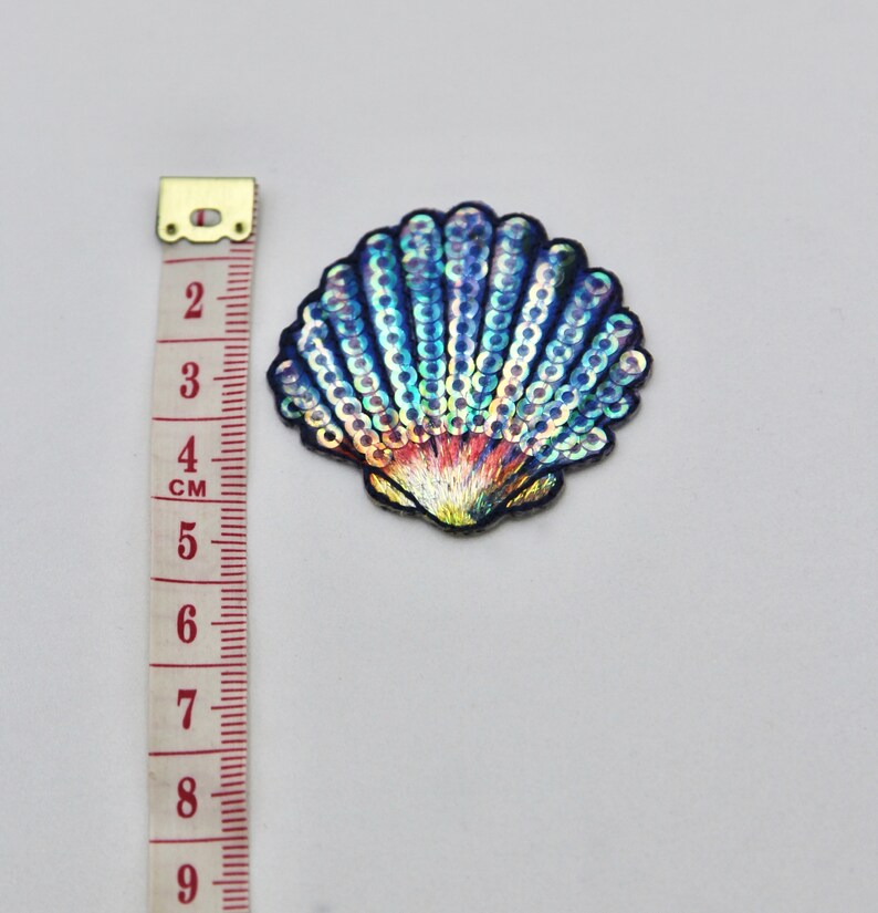 May include: A blue and iridescent seashell-shaped patch with sequins. The patch is about 4 cm wide.