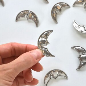 Puede incluir: Una colecci&oacute;n de dijes de plata en forma de media luna con un dise&ntilde;o de cara. Los dijes est&aacute;n esparcidos sobre una superficie blanca, con uno de ellos sostenido en una mano.