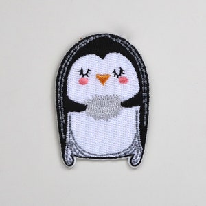 May include: A white and black embroidered penguin patch with a yellow beak and pink cheeks. The penguin has its eyes closed and is smiling.