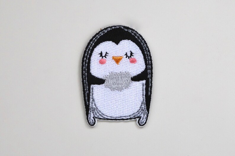 May include: A white and black embroidered penguin patch with a yellow beak and pink cheeks. The penguin has its eyes closed and is smiling.