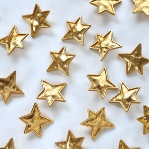 Puede incluir: Una colecci&oacute;n de adornos en forma de estrella de metal dorado esparcidos sobre un fondo blanco.