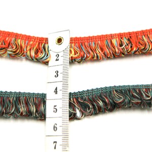 May include: Two rows of colorful fringe trim with a measuring tape showing 2 to 7 inches. The top row is red, orange, and blue. The bottom row is teal, red, and cream.