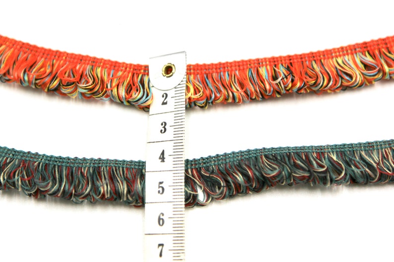 May include: Two rows of colorful fringe trim with a measuring tape showing 2 to 7 inches. The top row is red, orange, and blue. The bottom row is teal, red, and cream.