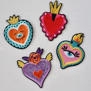 May include: Four embroidered iron-on patches in the shape of hearts with different designs. One heart is red with a crown, one is orange with a blue eye, one is pink with a blue eye and eyelashes, and one is purple with a red heart and wings.