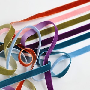 May include: Assortment of velvet ribbon in various colours, including teal, light blue, purple, green, pink, and orange. The ribbons are arranged in a loose pile on a white background.