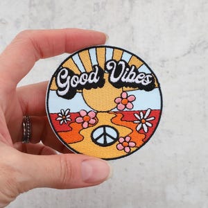 May include: A round embroidered patch featuring a sunset, flowers, and a peace sign. The text "Good Vibes" is in a retro font.