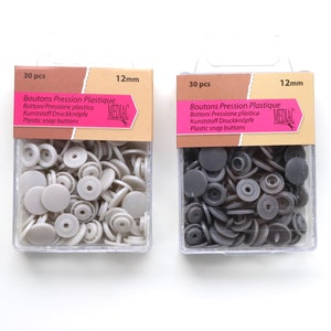 K&ouml;nnte beinhalten: Zwei Packungen mit je 30 Kunststoff-Druckkn&ouml;pfen, jeweils 12 mm im Durchmesser. Eine Packung enth&auml;lt graue Kn&ouml;pfe, die andere schwarze Kn&ouml;pfe. Die Packungen sind mit "Boutons Pression Plastique" und "MEDIAC" beschriftet.