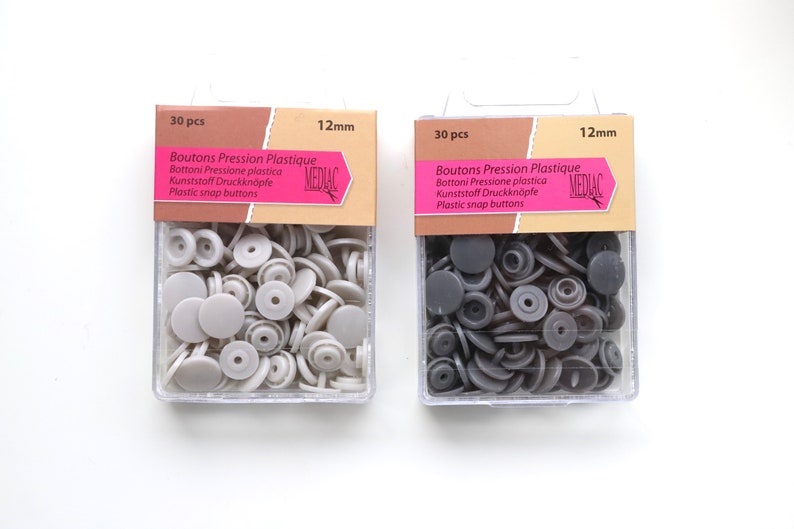 K&ouml;nnte beinhalten: Zwei Packungen mit je 30 Kunststoff-Druckkn&ouml;pfen, jeweils 12 mm im Durchmesser. Eine Packung enth&auml;lt graue Kn&ouml;pfe, die andere schwarze Kn&ouml;pfe. Die Packungen sind mit "Boutons Pression Plastique" und "MEDIAC" beschriftet.