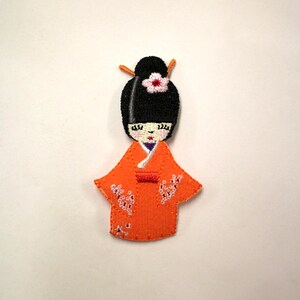 May include: Embroidered patch of a Japanese doll wearing a traditional orange kimono with white floral details. The doll has black hair, a white flower, and a purple sash.