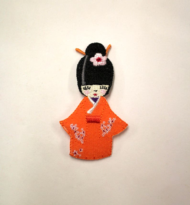 May include: Embroidered patch of a Japanese doll wearing a traditional orange kimono with white floral details. The doll has black hair, a white flower, and a purple sash.