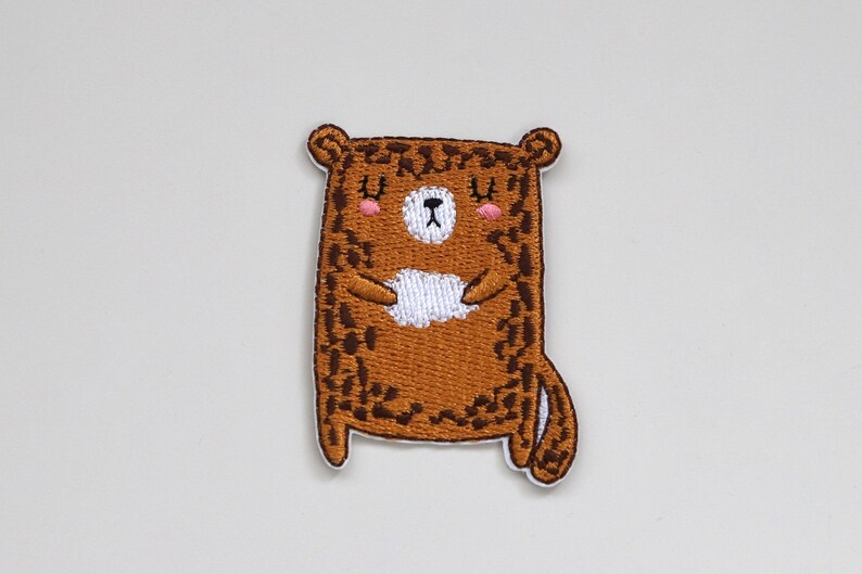 May include: Brown embroidered iron-on patch of a cartoon bear with a white belly and closed eyes. The bear has a brown and black leopard print pattern.