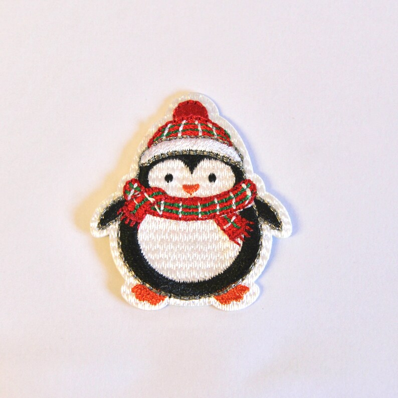 K&ouml;nnte beinhalten: Ein wei&szlig;er Filz-Pinguin mit einem rot-gr&uuml;nen karierten Schal und einer roten M&uuml;tze mit wei&szlig;em Rand. Der Pinguin ist mit schwarzen Augen, einem schwarzen Schnabel und schwarzen F&uuml;&szlig;en bestickt.