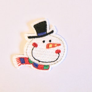 K&ouml;nnte beinhalten: Weisse Filz-Schneemann-Applikation mit einem schwarzen Zylinder, einer orangenen Nase und einem rot-gr&uuml;nen Schal.