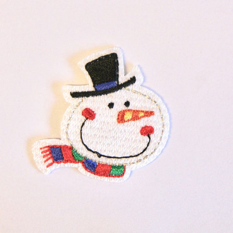 K&ouml;nnte beinhalten: Weisse Filz-Schneemann-Applikation mit einem schwarzen Zylinder, einer orangenen Nase und einem rot-gr&uuml;nen Schal.