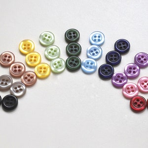 May include: A rainbow assortment of 24 plastic buttons, each with four holes. The buttons are arranged in a semi-circle, with the colours transitioning from light brown to dark red.