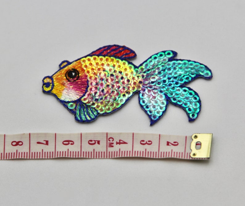 May include: A colorful iridescent fish-shaped sequin patch with blue, green, yellow, orange, and pink sequins. The patch is about 4 cm long and is perfect for adding a touch of whimsy to clothing or crafts.
