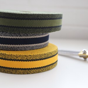 May include: Three rolls of striped fabric ribbon in yellow, navy blue, and green. The ribbon is stacked on a white surface with a pair of silver scissors.