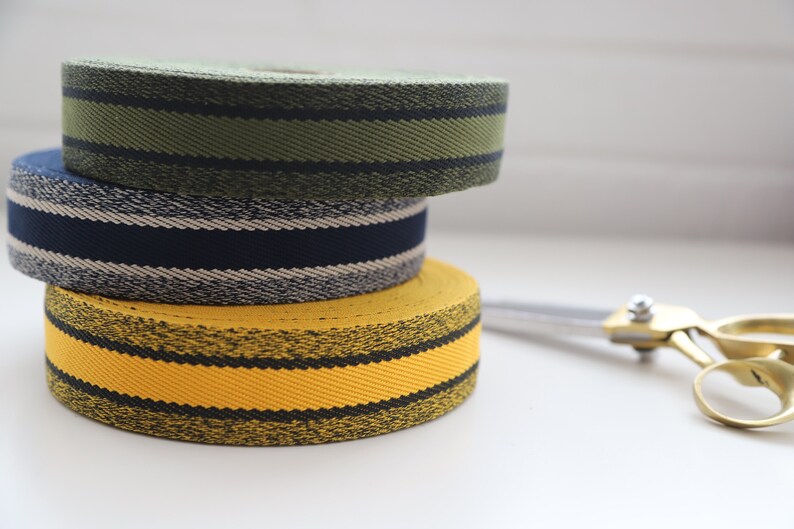 May include: Three rolls of striped fabric ribbon in yellow, navy blue, and green. The ribbon is stacked on a white surface with a pair of silver scissors.
