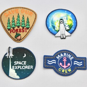 May include: Four embroidered patches with different designs. The first patch is tan with green trees and the text "SAVE OUR FOREST". The second patch is blue with a white space shuttle and the text "SPACE DREAM". The third patch is blue with a white space shuttle and the text "SPACE EXPLORER". The fourth patch is blue with a white anchor and the text "MARINE CREW".