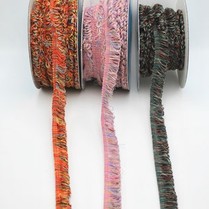May include: Three spools of colorful, fringed trim. The trim is orange, pink, and green. The trim is on spools with white labels.
