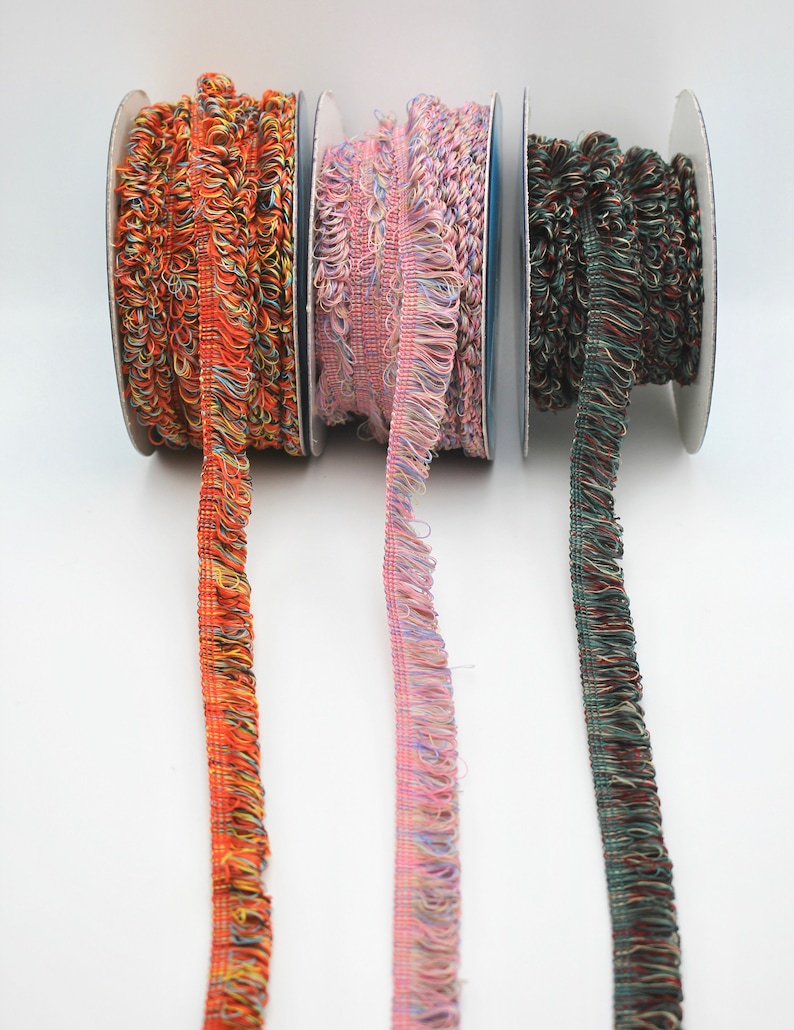 May include: Three spools of colorful, fringed trim. The trim is orange, pink, and green. The trim is on spools with white labels.