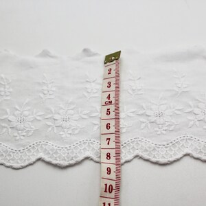 Peut inclure: Bordure en dentelle de coton blanche avec un bord festonn&eacute; et un motif floral brod&eacute;. La bordure est mesur&eacute;e avec une r&egrave;gle indiquant 11 cm.