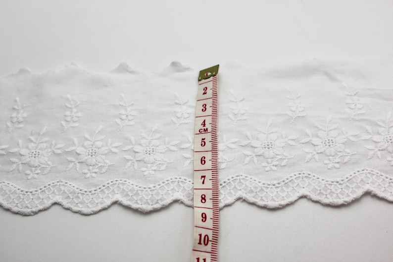 Peut inclure: Bordure en dentelle de coton blanche avec un bord festonn&eacute; et un motif floral brod&eacute;. La bordure est mesur&eacute;e avec une r&egrave;gle indiquant 11 cm.