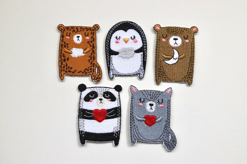 May include: Five embroidered iron-on patches featuring cute animal designs. The patches include a brown bear holding a moon, a penguin, a brown bear holding a heart, a panda holding a heart, and a gray cat holding a heart.